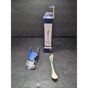 May include: A small, decorative spoon with a green and yellow design, next to its blue box. The box has a label with text and an illustration of the spoon. The spoon is on a dark surface.