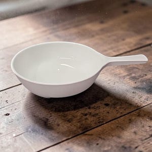 May include: A white serving bowl with a handle, set against a wooden surface. The bowl has a smooth, rounded shape and a handle extending from one side. The handle is rectangular and flat.