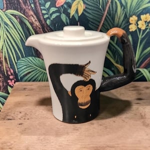 May include: White ceramic teapot with a black monkey design. The monkey's arm forms the handle, with a brown hand. The teapot has a white lid and a spout for pouring. The design is whimsical and decorative.