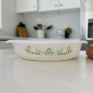 May include: Off-white oval baking dish with green floral design. The dish has two handles and is set on a white surface. The kitchen background includes cabinets and utensils.