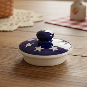 May include: A round ceramic lid with a glossy dark blue finish featuring a pattern of white stars. The lid has a small, rounded knob handle at the center and a solid white base rim designed to fit securely onto a corresponding container vessel.