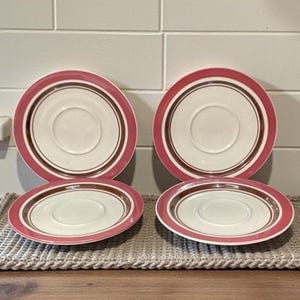May include: Four vintage saucers with a white center, a brown band, and a pink outer rim. The saucers are arranged in two stacks of two on a woven placemat. The saucers are made of ceramic.