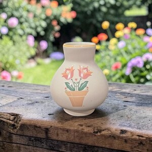 Könnte beinhalten: Kleine, cremefarbene Keramikvase mit floralem Design. Die Vase zeigt eine gemalte Illustration von zwei orangefarbenen Tulpen in einem Terrakotta-Topf. Der Rand der Vase hat einen dünnen, gelben Rand. Die Vase steht auf einer Holzoberfläche.
