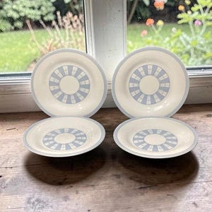 May include: Four off-white ceramic plates with a light blue geometric design. The plates have a circular pattern in the center and a light blue border. The plates are arranged on a wooden surface near a window.