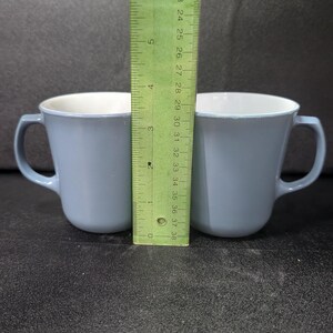 Two light blue coffee mugs placed side by side with a green ruler standing upright between them against a dark surface.