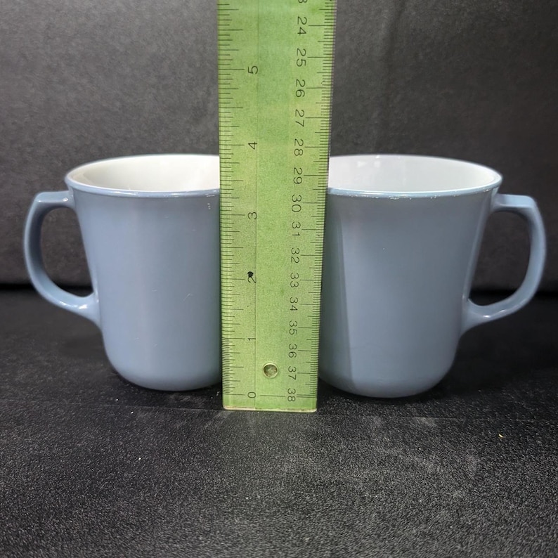 Two light blue coffee mugs placed side by side with a green ruler standing upright between them against a dark surface.