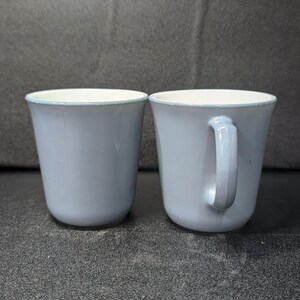Two light blue coffee mugs positioned side by side, showing one mug viewed from the front and the other from the side with its handle.