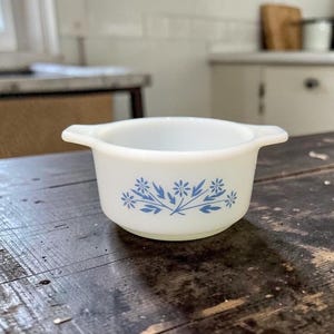 May include: A white, small, round bowl with two handles. The bowl features a blue floral design around the exterior. The bowl is sitting on a dark wooden surface. The bowl is likely used for serving or baking.