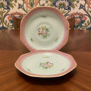 May include: Two octagonal ceramic saucers featuring a pale mint green center with a central floral bouquet of pink, yellow, and purple flowers. The wide rim is decorated with a textured pink pattern and a thin gold edge, with small floral sprigs.
