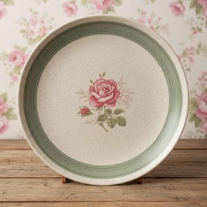 May include: A round ceramic dinner plate with a speckled cream-colored base. The plate features a central illustration of a pink rose with green leaves and a wide, sage green band encircling the inner rim, creating a simple and clean aesthetic.