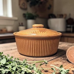 May include: A covered, round, tan ceramic casserole dish with vertical ribbed detailing. The lid has a small handle and horizontal ribbed detailing. The dish sits on a wooden surface with green herbs.