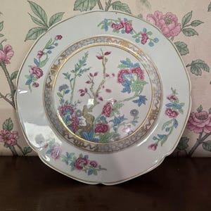 May include: A white porcelain plate with a scalloped edge, featuring a central floral design in pink, blue, and green, framed by a gold border. Additional floral patterns adorn the plate's rim, creating a vintage aesthetic.