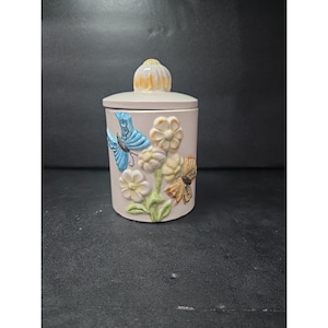 Puede incluir: Un tarro de cerámica con tapa, con mariposas y flores en relieve. El tarro es de color beige claro, con detalles azules, amarillos y marrones en las mariposas y flores. La tapa tiene un diseño floral amarillo y blanco.