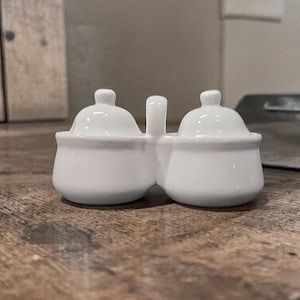 Op de afbeelding: Een witte keramische zout- en peperstrooier set met twee verbonden compartimenten, elk met een klein deksel en een handvat ertussen. De set staat op een houten oppervlak.