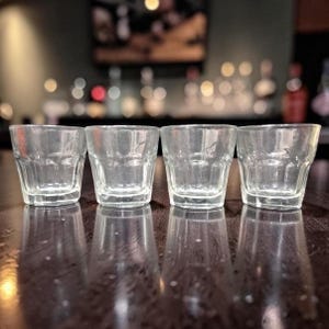 May include: Four clear, empty shot glasses are arranged in a row on a dark wooden surface. The glasses have a thick, square base and a slightly flared rim. The background is blurred, suggesting a bar setting.