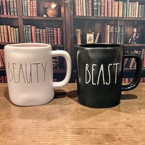 May include: Two ceramic mugs, one white and one black, are displayed side-by-side. The white mug has the word "BEAUTY" in black lettering, and the black mug has the word "BEAST" in white lettering. Both mugs have a rounded shape and a large handle.