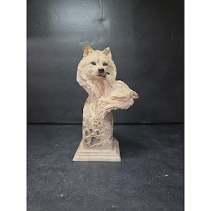 May include: A detailed sculpture of two wolves, one embracing the other. The wolves are depicted in a light beige color with textured fur, mounted on a square base. The sculpture is set against a dark background.