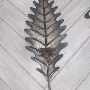 Puede incluir: Una lámpara de pared de metal con forma de hoja con un portavelas. La hoja es texturizada y tiene un acabado marrón oscuro.