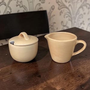 May include: A cream-coloured ceramic sugar bowl with a lid and a small handle, beside a matching creamer with a handle and spout. Both items are on a dark wooden surface. The set is likely for serving coffee or tea.