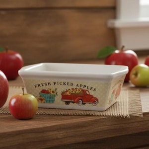 May include: Rectangular ceramic dish with the text "Fresh Picked Apples". The dish has a cream-colored background with illustrations of a blue basket of sunflowers and a red truck filled with apples. Red and green apples are scattered around the dish.