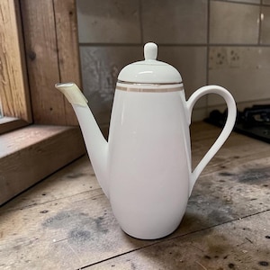May include: A white ceramic coffee pot with a gold band around the lid and a curved handle. The spout has a piece of tape on it. The pot is on a wooden surface.