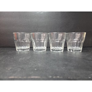 May include: Four clear, small, square-shaped shot glasses. Each glass has a thick, ridged base and a straight, slightly flared rim. The glasses are empty and arranged in a row against a dark background.