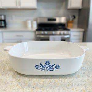Puede incluir: Una fuente de horno rectangular de cerámica blanca con asas laterales integradas. El exterior presenta un patrón floral azul centrado que consiste en tres flores estilizadas con hojas. El interior es liso, blanco y profundo con esquinas redondeadas.