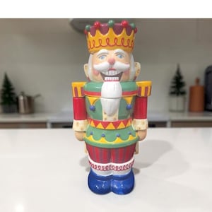 May include: A colorful, decorative nutcracker figurine in the shape of a king. The figurine features a crown, a smiling face, and a uniform with red, green, yellow, and blue accents. The base is blue with a decorative swirl design.