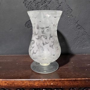 Puede incluir: Un jarrón de vidrio esmerilado con una parte superior acampanada y una base redonda. El jarrón presenta un patrón grabado de formas abstractas arremolinadas. El jarrón está sobre una superficie de madera oscura, con un fondo gris oscuro.