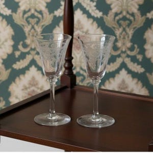 May include: Two clear, stemmed glasses with etched floral designs. The glasses are on a dark brown wooden surface, with a patterned wallpaper background. The glasses have a classic, elegant appearance.