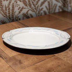 May include: An oval, white serving platter with a silver rim and a delicate blue floral pattern. The platter is set on a wooden surface, showcasing its elegant design. This dish is perfect for serving appetizers or desserts.