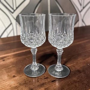 May include: Two clear crystal wine glasses with a detailed, textured pattern on the bowl and stem. The glasses are displayed on a dark wooden surface, showcasing their elegant design. Perfect for serving wine.