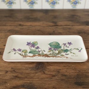 May include: Rectangular white ceramic serving dish with a floral design featuring purple violets, green leaves, and brown roots. The dish has a thin, light brown border and is set on a wooden surface.