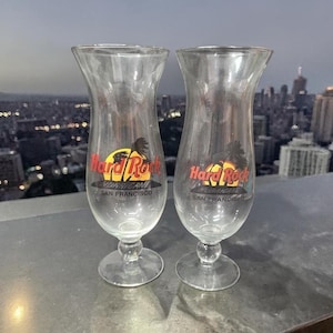 Puede incluir: Dos vasos de vidrio transparente Hurricane con el logotipo de Hard Rock Cafe con un sol amarillo, palmeras y el texto "Hurricane San Francisco". Los vasos tienen forma acampanada y un tallo.