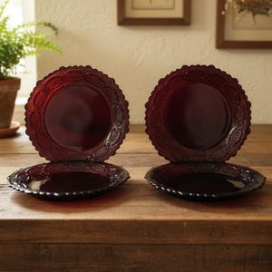 May include: Two pairs of deep red glass plates featuring a scalloped edge and a textured, embossed pattern along the rim. Each set consists of one plate standing upright and one plate lying flat, showcasing the intricate, repeating geometric design.