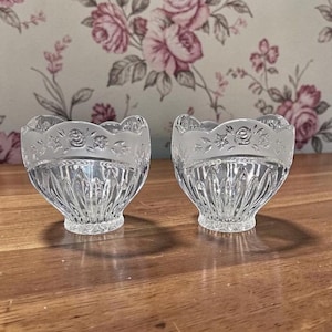 May include: Two clear glass bowls with a scalloped rim featuring a floral design. The bowls have a textured, cut-glass pattern on the lower half and sit on a wooden surface. The background is a floral wallpaper.