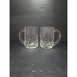 May include: Two clear glass beer mugs with textured sides and handles. The mugs are empty and sit on a dark surface. The mugs are designed for holding beer or other beverages.