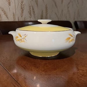 Puede incluir: Un cuenco de servir vintage blanco y amarillo con tapa. La tapa y la base son amarillas, mientras que el cuerpo es blanco con diseños de espigas de trigo. El cuenco tiene dos asas y un pequeño pomo en la tapa.