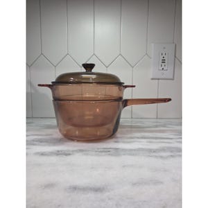 Puede incluir: Una cacerola de vidrio ámbar vintage con tapa y asa. La cacerola está hecha de vidrio transparente, lo que permite ver el contenido. La tapa tiene un pomo marrón oscuro. La cacerola está sobre una encimera de mármol.