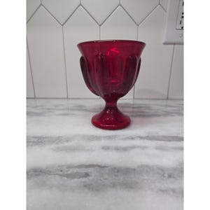 May include: A ruby red glass goblet with a decorative, fluted design. The goblet has a rounded bowl and a short, stemmed base. The glass is transparent, allowing light to pass through. The goblet is likely used for serving beverages.
