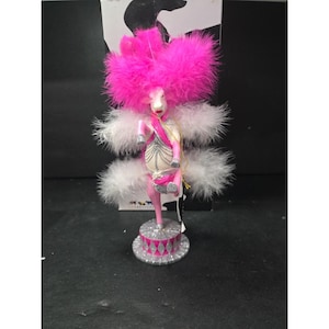 May include: A whimsical figurine of a pink sheep with a large pink feather headdress and white feather wings. The sheep wears a pink sash and holds a small pink purse. It stands on a silver and pink circus-style base.