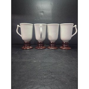 May include: Four white ceramic mugs with brown pedestal bases and curved handles. The mugs are arranged in a row, showcasing their design. The mugs are suitable for serving hot beverages.