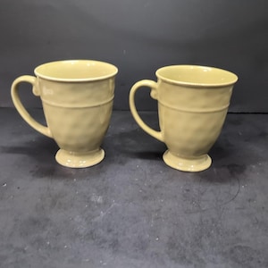 Puede incluir: Dos tazas de cerámica con un esmalte texturizado de color marrón claro. Las tazas tienen un cuerpo redondeado y una base acampanada. Tienen un asa en un lado.