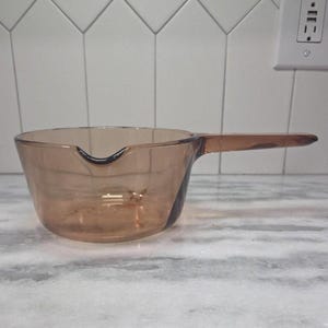 Könnte beinhalten: Ein durchsichtiger, bernsteinfarbener Glas-Kochtopf mit langem Griff und Ausguss. Der transparente Glas ermöglicht die Sichtbarkeit des Inhalts. Der Griff erstreckt sich horizontal von der Seite und bietet einen sicheren Griff für eine einfache Handhabung. Für die Küche.