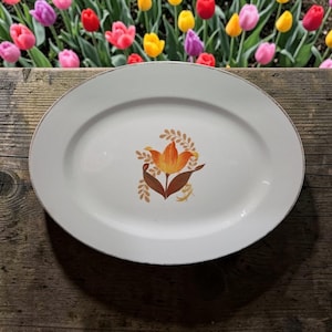 Könnte beinhalten: Ovale weiße Servierplatte mit goldenem Rand und floralem Design in der Mitte. Die Blume ist orange und braun mit grünen Akzenten. Die Platte steht auf einer Holzoberfläche.