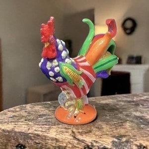 May include: A vibrant ceramic rooster ornament. It features a red comb, a purple body with white spots, and a red and white striped chest. The rooster has green, orange, and yellow details, and stands on an orange base. A decorative item.