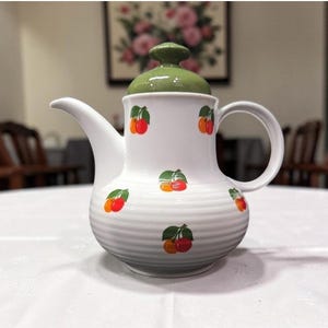 May include: White ceramic teapot with a green lid and handle, adorned with red and orange cherry illustrations. The teapot has a curved spout and a ribbed design around the base. A decorative kitchenware item.