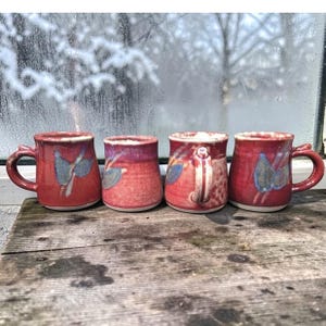 Puede incluir: Cuatro tazas de cerámica hechas a mano de diferentes tamaños, cada una con un esmalte rojo y diseños de hojas azules. Las tazas tienen formas de asa únicas y se exhiben sobre una superficie de madera desgastada, con una ventana nevada al fondo.