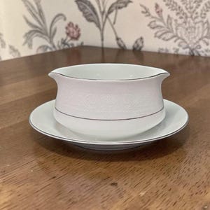 May include: A white gravy boat with a matching saucer. The boat has a curved spout and a decorative embossed pattern. Both pieces have a silver trim. The set is on a wooden surface.