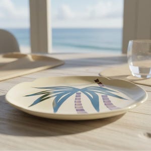 May include: A round, cream-colored plate featuring a stylized graphic of a palm tree with blue fronds and a purple segmented trunk. The plate has a smooth, slightly scalloped edge and is positioned on a light-colored surface near a clear glass.